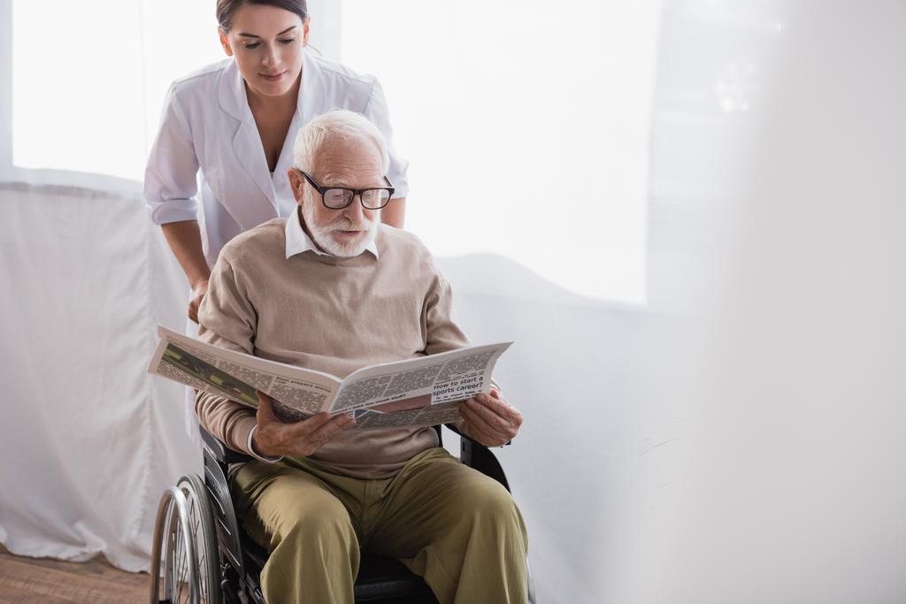 車椅子で新聞を読む高齢障害者の近くの老人看護師 - 写真・画像