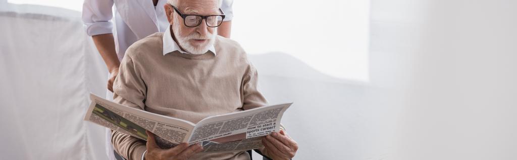 老人ホームのソーシャルワーカーの近くで新聞を読む眼鏡の男バナー - 写真・画像