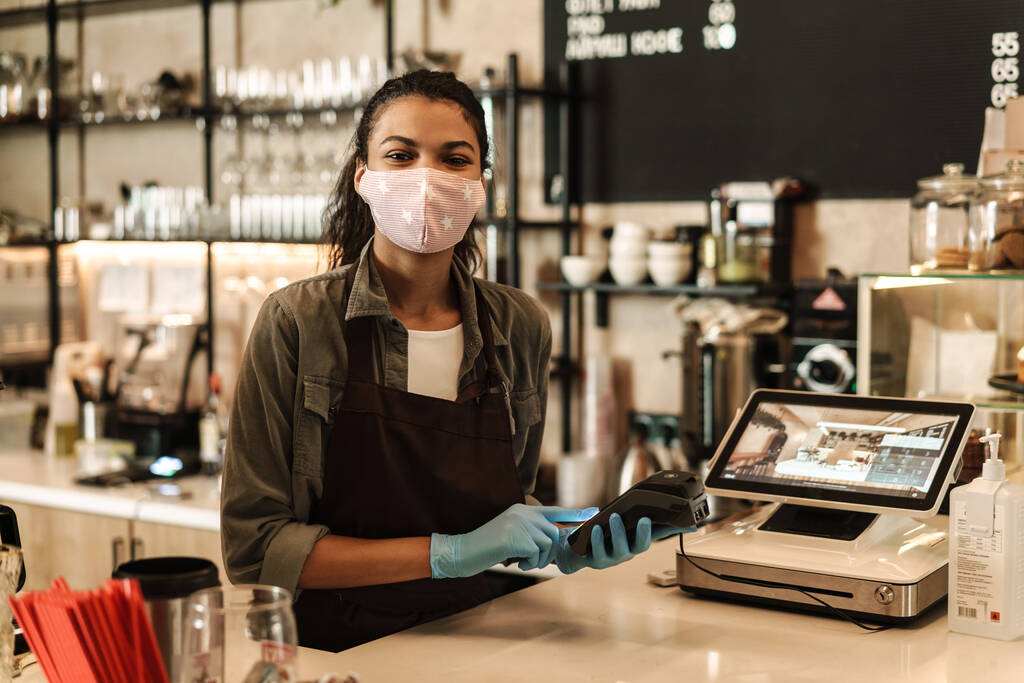 Mulher barista vestindo máscara facial médica segurando terminal sem dinheiro para pagar a ordem no balcão - Foto, Imagem