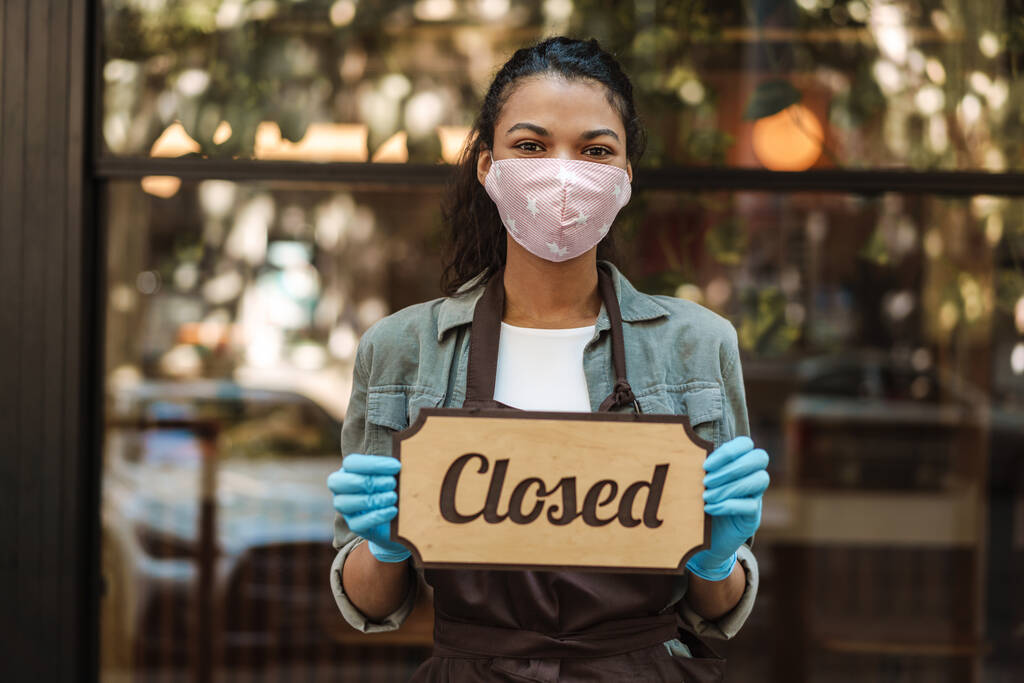 Жінка в медичній масці для обличчя тримає закритий знак, стоячи перед кафе
 - Фото, зображення