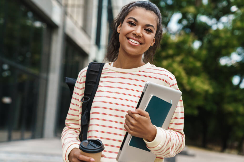 街の通りでノートパソコンと歩いている間に楽しいアフリカ系アメリカ人の女の子がコーヒーを飲むイメージ - 写真・画像