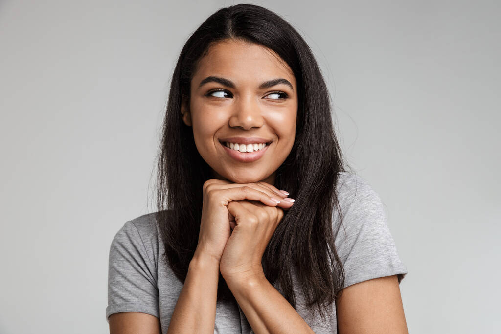 Portrait of a happy smiling young african woman with long dark hair wearing casual clothes standing isolated over gray background, looking away - Photo, Image
