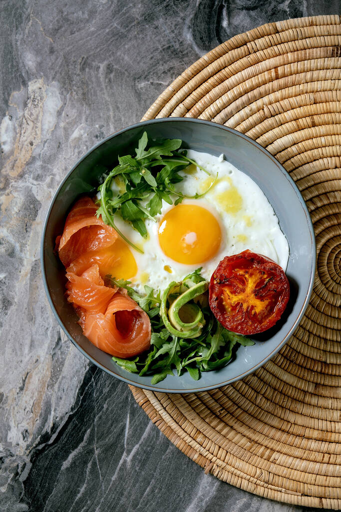 Zdrowa miska śniadaniowa ze smażonymi jajami, łososiem, awokado, grillowanym pomidorem i sałatką serwującą chleb na słomkowej serwetce na ciemnym marmurowym tle. Płaskie ułożenie, przestrzeń kopiowania - Zdjęcie, obraz