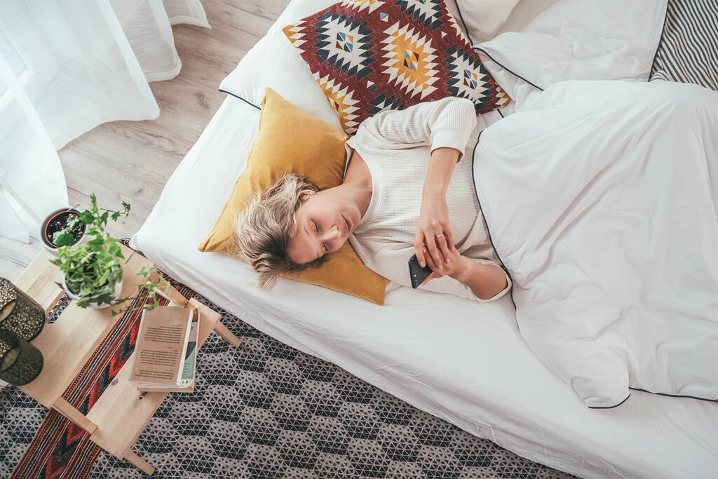 Jonge blonde vrouw gekleed pijama liggend in het gezellige bed onder de deken en surfen op een smartphone internet. Zoete gezellige huis en draadloze technologie concept imago. - Foto, afbeelding