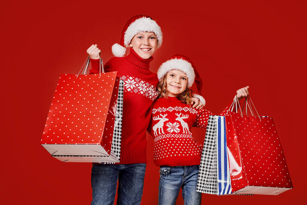 Enfants mignons en chapeaux rouges Santa, chandails tenant des sacs à provisions sur fond rouge - Photo, image