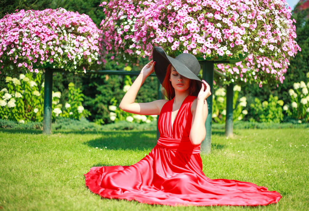 jong aantrekkelijk meisje in een lange rode jurk met een zwarte hoed, in een bloementuin op het gazon - Foto, afbeelding