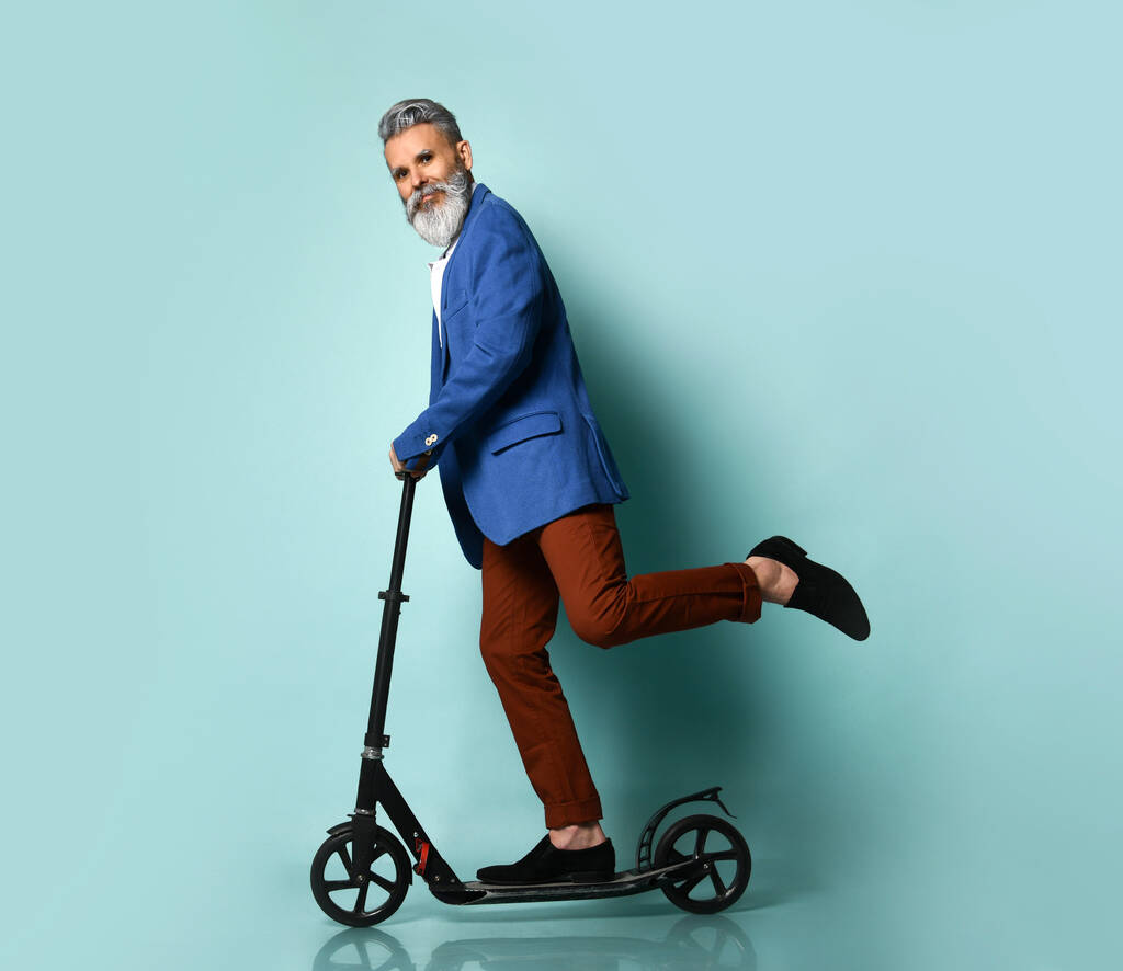 Beyaz gömlekli, ceketli, kahverengi pantolonlu, mokasen giyen gri saçlı yaşlı bir adam. Siyah scooter 'a binmek ve mavi arka planda yan poz vermek - Fotoğraf, Görsel