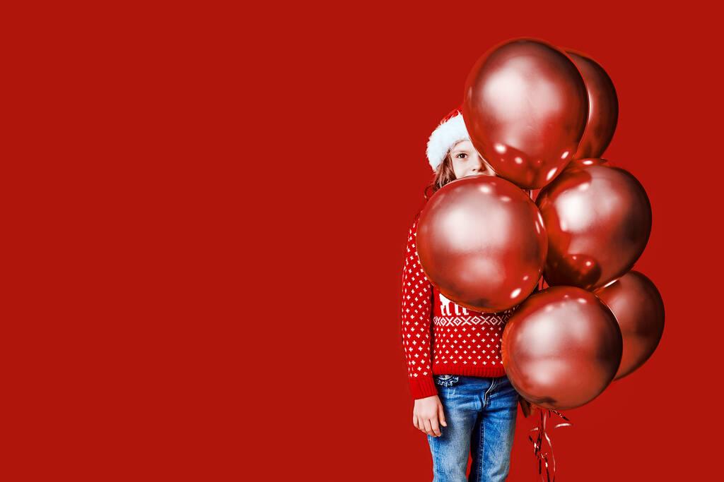Χαριτωμένο παιδί σε κόκκινο καπέλο Σάντα και πουλόβερ κρύβονται πίσω από μπαλόνια σε κόκκινο φόντο - Φωτογραφία, εικόνα