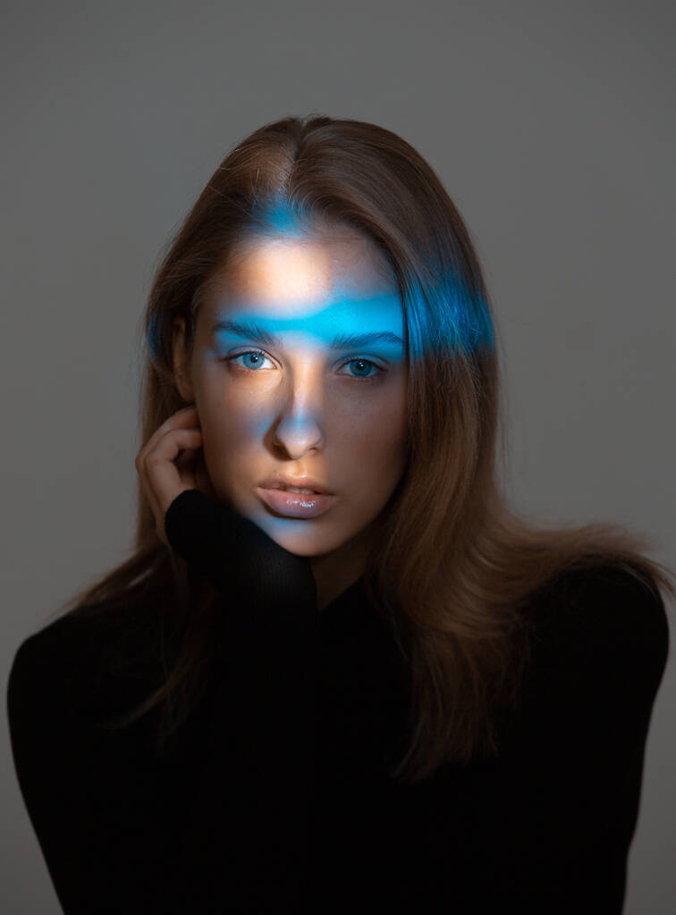 Δημιουργικό πορτρέτο της κομψής νεαρής γυναίκας με μπλε νέον φως στο πρόσωπο και τέλειο δέρμα σε γκρι φόντο - Φωτογραφία, εικόνα