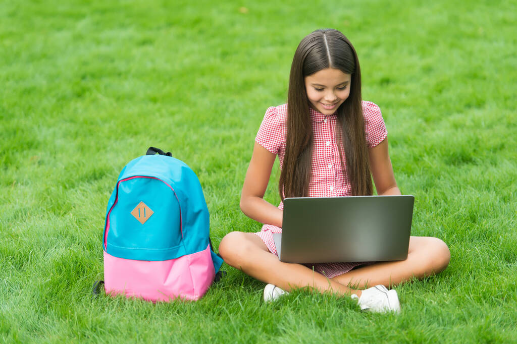 幸せな子供がノートパソコンで働いてる。オンライン教育です。学校に戻って。10代の女の子は公園の緑の芝生でコンピューターを使う。リュックとノートを持った子供。現代の生活における新しい技術です。勉強しろ - 写真・画像