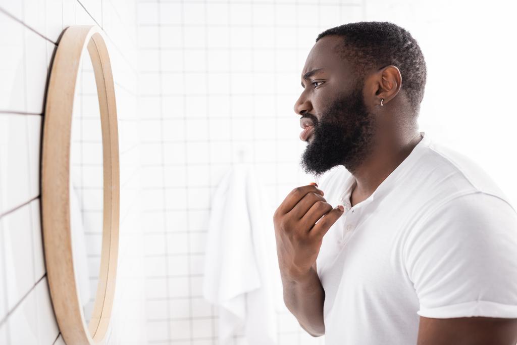 πλαϊνή όψη δυστυχισμένου αφροαμερικανού που κοιτάζει τα γένια στον καθρέφτη - Φωτογραφία, εικόνα