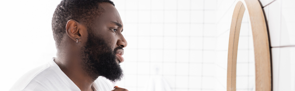 plano panorámico de hombre afro-americano infeliz mirando la barba en el espejo - Foto, Imagen