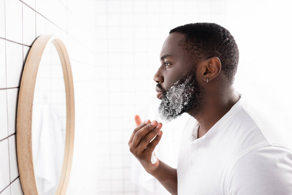 πλαϊνή όψη Αφρο-Αμερικανού που απλώνει αφρό ξυρίσματος και κοιτά στον καθρέφτη - Φωτογραφία, εικόνα