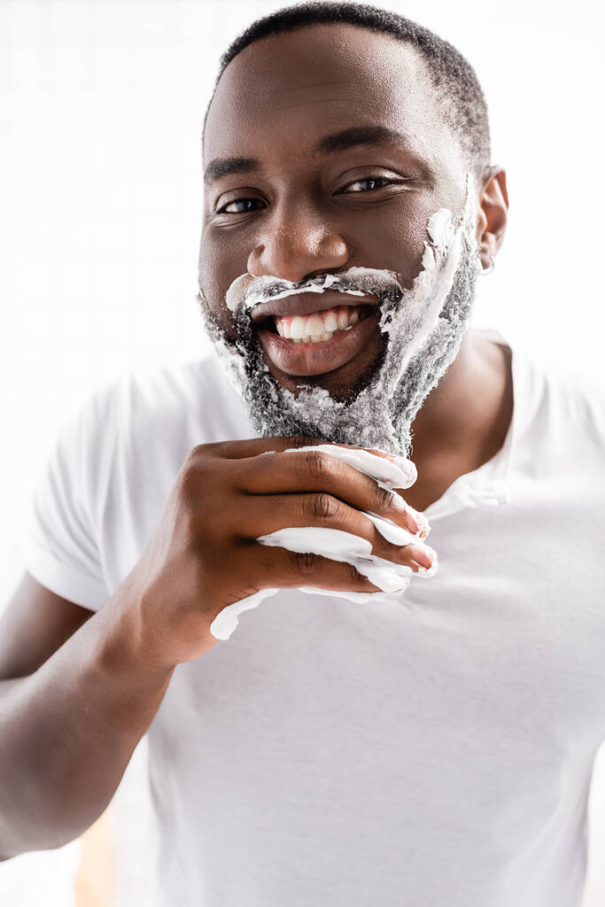 hombre afro-americano sonriente con espuma de afeitar en la cara mirando la cámara - Foto, Imagen