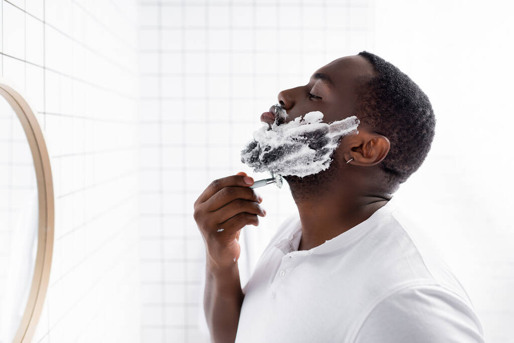 Afro-Amerikan bir adamın tıraş olurken sakalını tıraş edişinin yan görüntüsü.  - Fotoğraf, Görsel