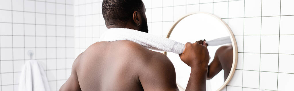 panoramisch schot van afro-Amerikaanse man op zoek in spiegel met handdoek achter de nek  - Foto, afbeelding