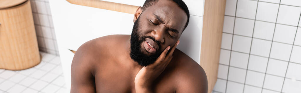 panoramic shot of afro-american man sitting on bathroom floor and grimacing from toothache - Photo, Image