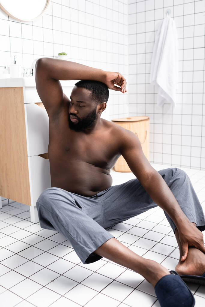 afro-american man with hand up sitting on floor and smelling armpit - Photo, Image