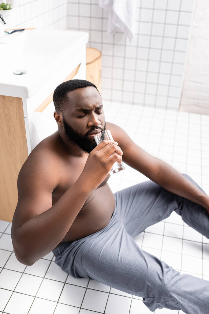 homme afro-américain assis sur le sol de la salle de bain et l'eau potable - Photo, image