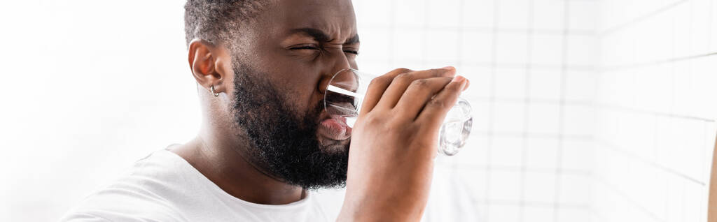 панорамный снимок афро-американского человека, гримасирующего и пьющего воду - Фото, изображение