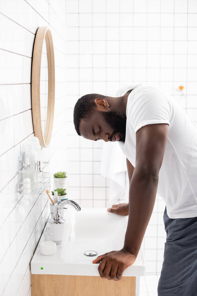 Müder afroamerikanischer Mann mit geschlossenen Augen lehnt sich an Waschbecken  - Foto, Bild