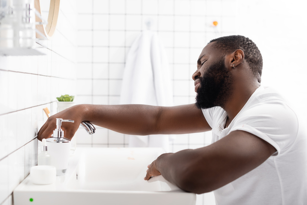 Müder afroamerikanischer Mann lehnt am Waschbecken und versucht, den Wasserhahn aufzudrehen - Foto, Bild