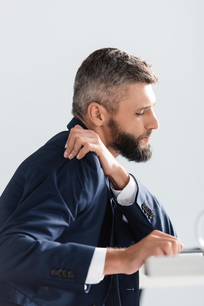 Vista lateral do homem de negócios em terno sentindo dor no ombro perto de primeiro plano borrado no escritório  - Foto, Imagem