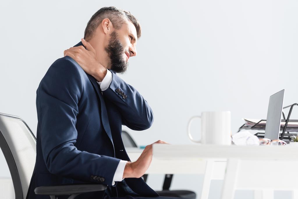Geschäftsmann in offizieller Kleidung leidet unter Nackenschmerzen in der Nähe von Laptop und Tasse im verschwommenen Vordergrund im Büro  - Foto, Bild