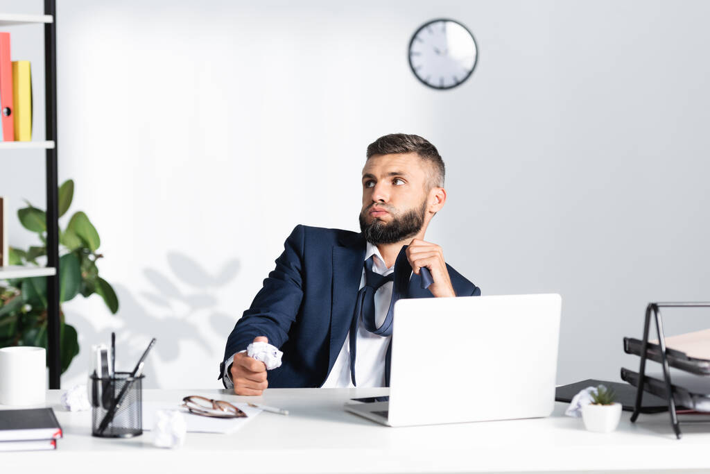 Pensive бізнесмен торкається краватки і тримає згорнутий папір біля ноутбука і канцелярських приладів на розмитому передньому плані в офісі
  - Фото, зображення