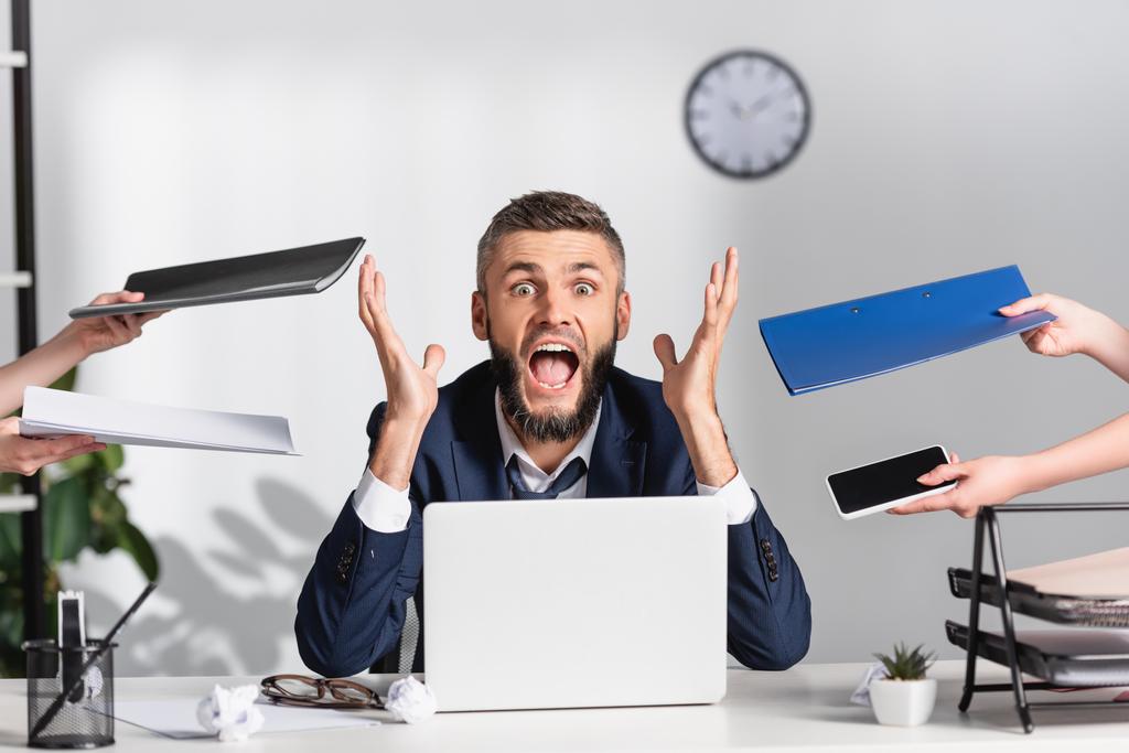 Gestresste zakenman schreeuwen in de buurt van collega 's met papieren mappen en gadgets in het kantoor  - Foto, afbeelding