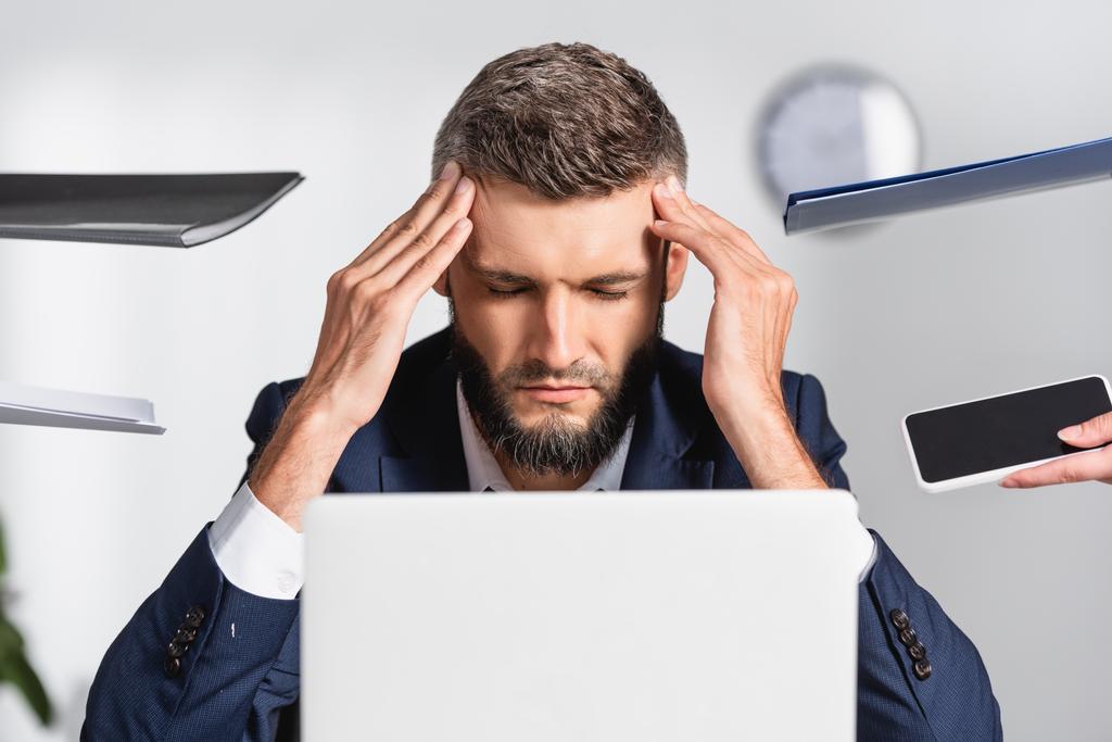 Unavený podnikatel dotýkající se hlavy v blízkosti papírových složek a gadgets na rozmazané popředí v úřadu - Fotografie, Obrázek