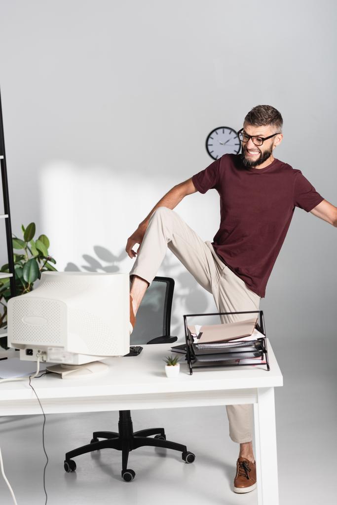 Vihainen liikemies potkimassa tietokonenäyttö lähellä paperitavarat toimistossa  - Valokuva, kuva