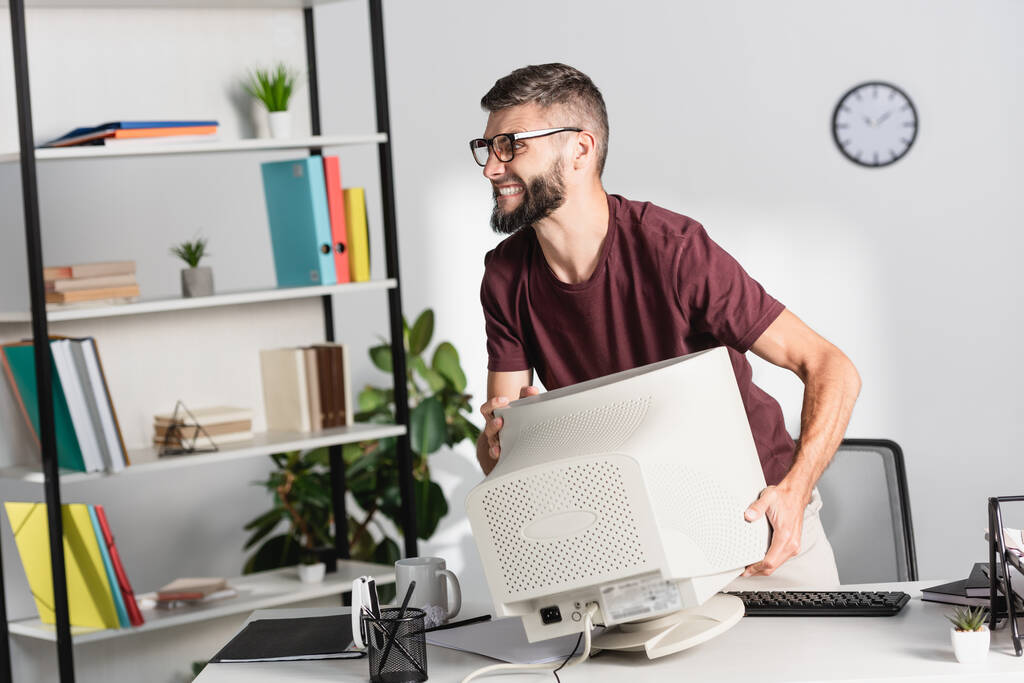 Θυμωμένος επιχειρηματίας κρατώντας οθόνη υπολογιστή κατά τη διάρκεια νευρικού κλονισμού στο γραφείο  - Φωτογραφία, εικόνα