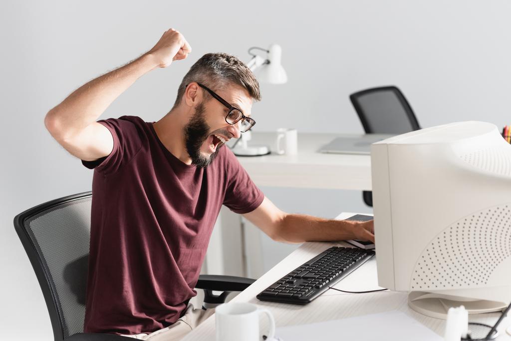 Schreiender Geschäftsmann sitzt bei Nervenzusammenbruch im Büro neben Computer im verschwommenen Vordergrund  - Foto, Bild
