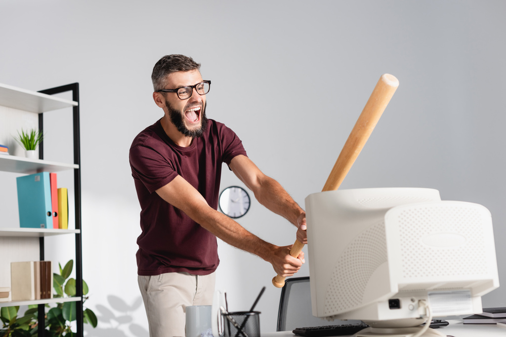Screaming businessman beating computer monitor with baseball bat on blurred foreground on office table  - Photo, Image