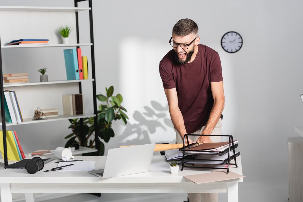 Homem de negócios irritado batendo laptop com bastão de beisebol perto de artigos de papelaria na mesa de escritório  - Foto, Imagem