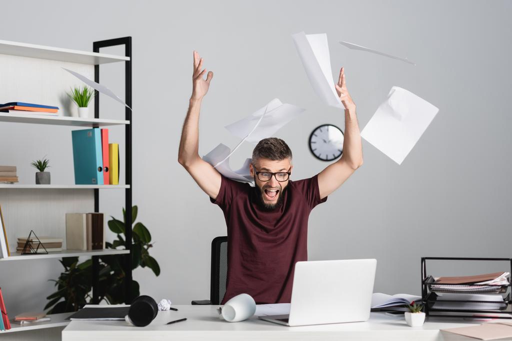 Stres podnikatel křičí při házení papíry v blízkosti notebooku na kancelářský stůl  - Fotografie, Obrázek