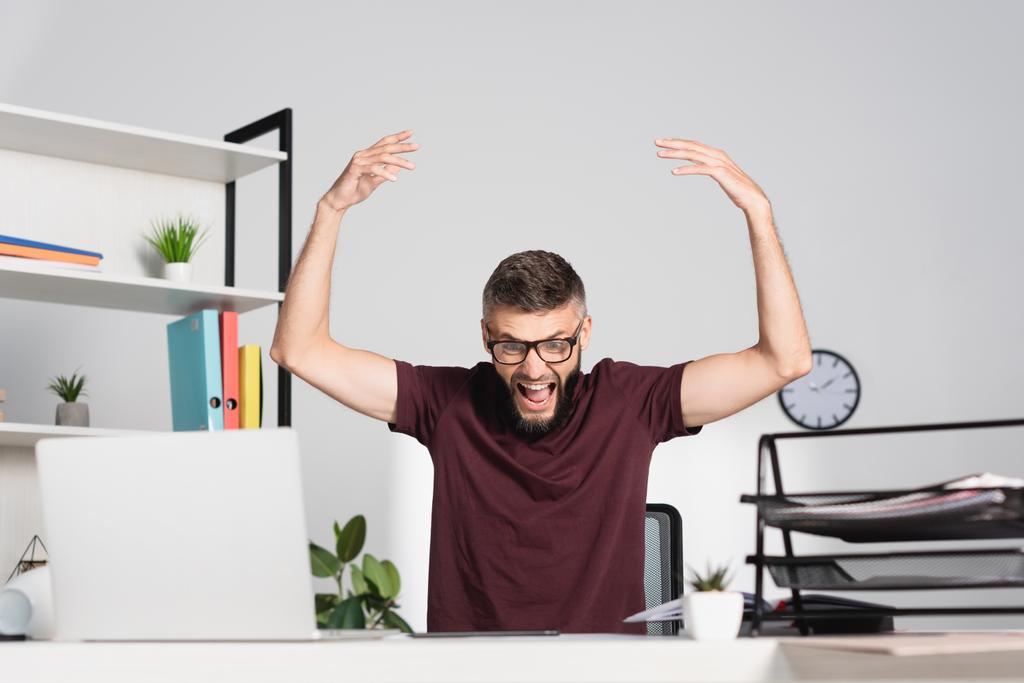 Hombre de negocios enojado gritando cerca de la computadora portátil y artículos de papelería en primer plano borroso en la oficina  - Foto, Imagen