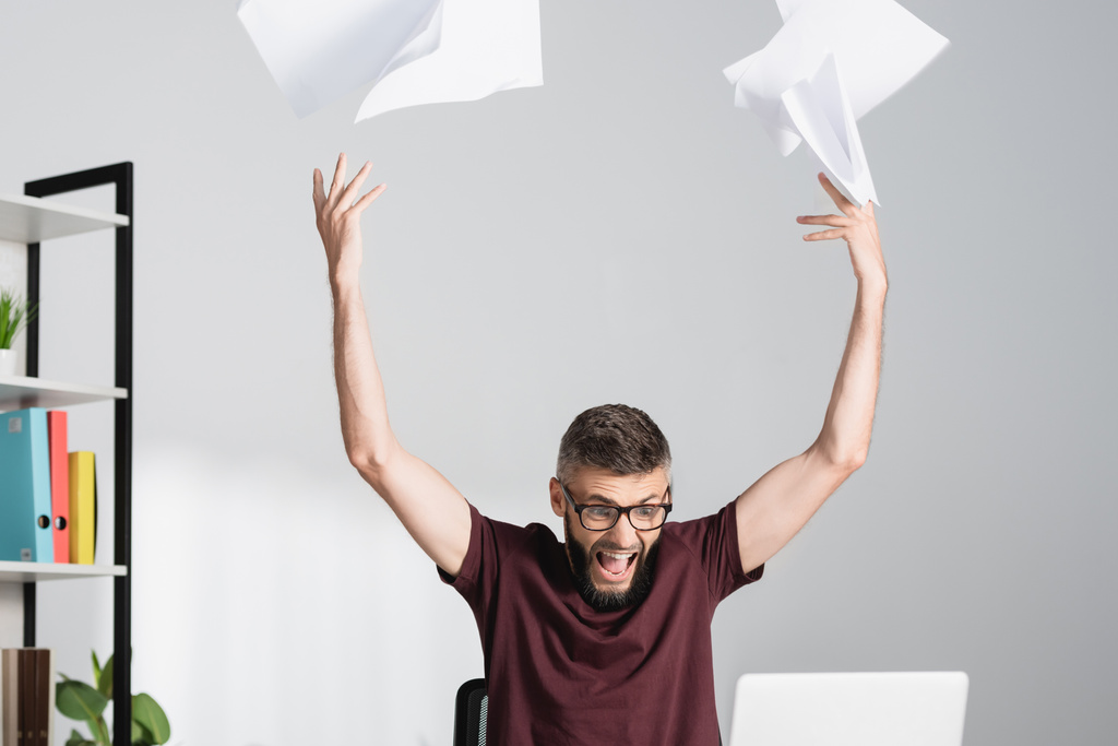 Jännittynyt ja huutava liikemies heittää asiakirjoja läppärin lähelle hämärtyneellä etualalla toimistossa  - Valokuva, kuva