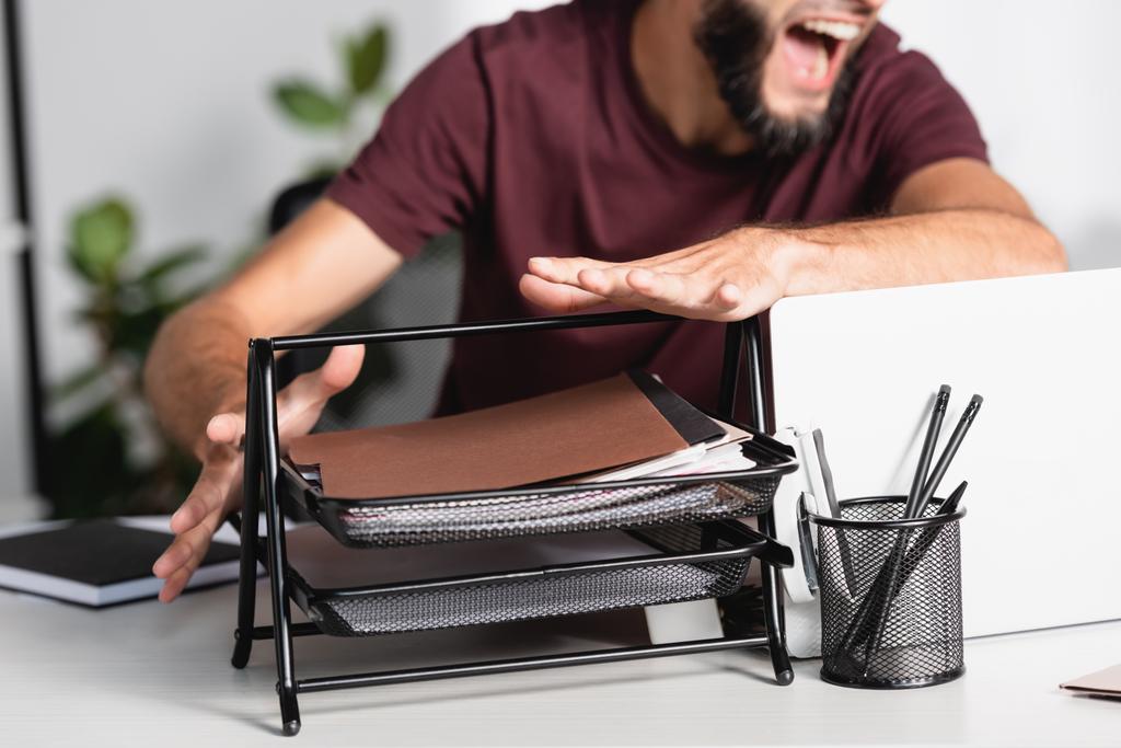 Обрезанный вид кричащего бизнесмена, бросающего канцелярские принадлежности и ноутбук со стола  - Фото, изображение