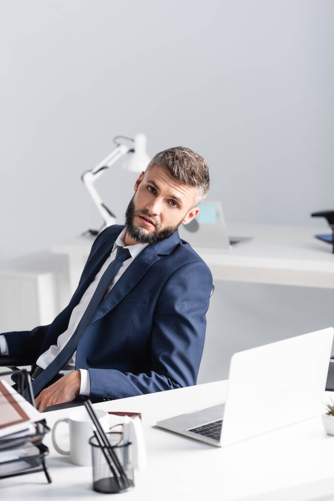 Бизнесмен в костюме смотрит в камеру рядом с ноутбуком и канцелярскими принадлежностями на размытом переднем плане в офисе  - Фото, изображение