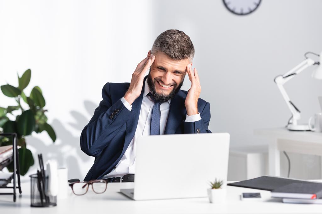 Uomo d'affari che soffre di mal di testa vicino a laptop e documenti sul tavolo da lavoro  - Foto, immagini