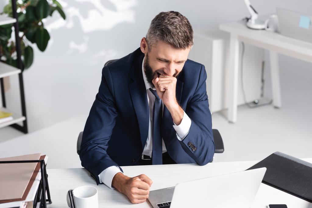 Επιχειρηματίας χασμουριέται ενώ εργάζεται κοντά σε φορητό υπολογιστή και χαρτιά στο τραπέζι  - Φωτογραφία, εικόνα