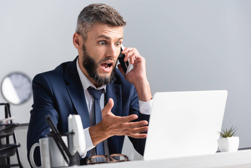 Emocionado hombre de negocios hablando en el teléfono inteligente y apuntando a la computadora portátil en primer plano borrosa  - Foto, imagen