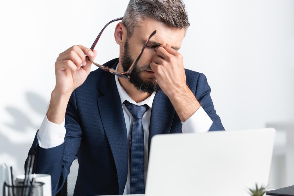 Empresário cansado segurando óculos enquanto trabalhava perto do laptop em primeiro plano desfocado  - Foto, Imagem