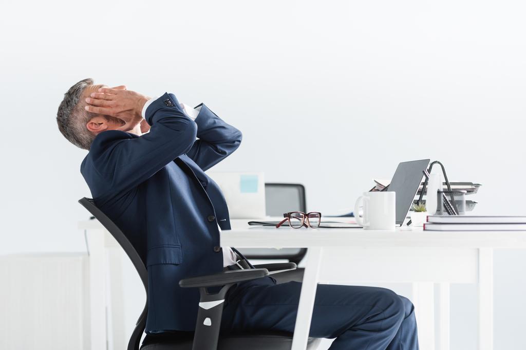 Homme d'affaires fatigué touchant le visage près d'un ordinateur portable et des lunettes sur le premier plan flou au bureau   - Photo, image