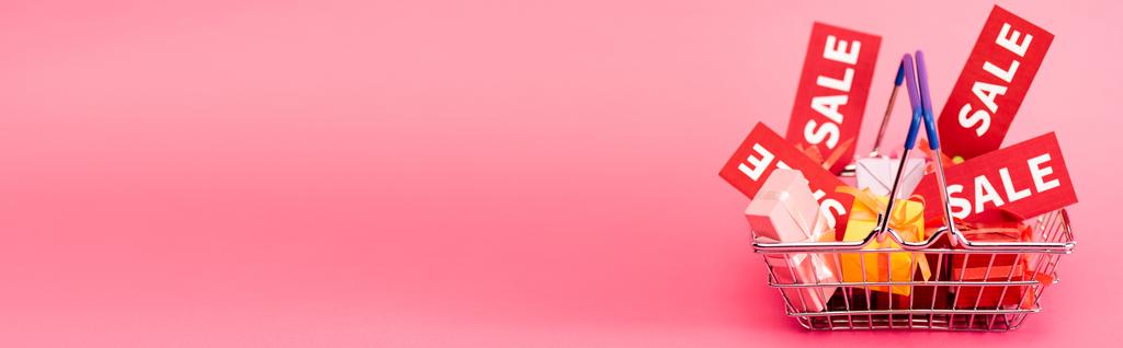panoramisch gewas van boodschappenmandje met cadeautjes en rode sale tags op roze  - Foto, afbeelding