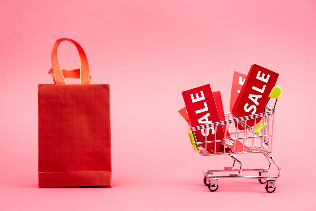 etiquetas vermelhas com letras de venda no carrinho de compras perto de saco de papel em rosa  - Foto, Imagem