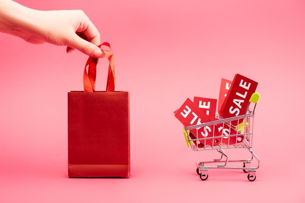 обрезанный вид женской руки касаясь сумки рядом с корзиной с этикетками продажи на розовый - Фото, изображение