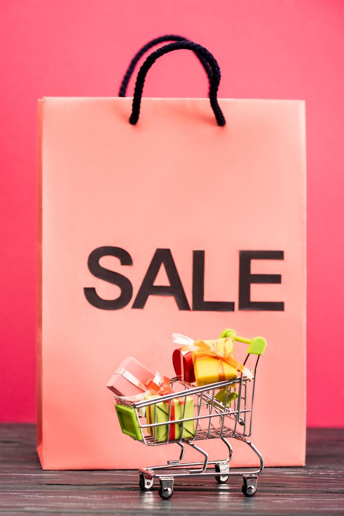 селективный фокус игрушечной тележки с подарками возле бумажного пакета с надписью продажи на розовой, черной пятничной концепции  - Фото, изображение